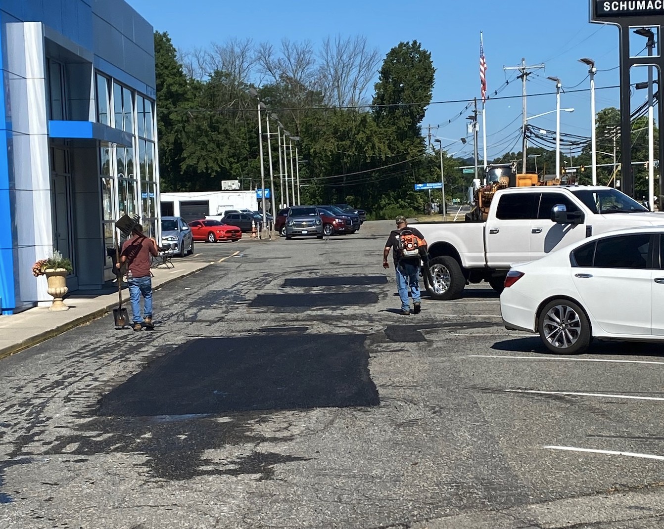 Two people with backpacks walk through a parking lot with various parked vehicles, including white and gray cars, near a building with blue trim on a sunny day. Trees and utility poles are visible in the background.
